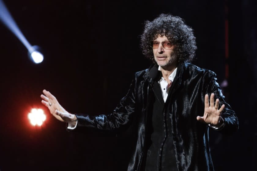 A man with bushy, curly hair in a shiny black suit speaks onstage