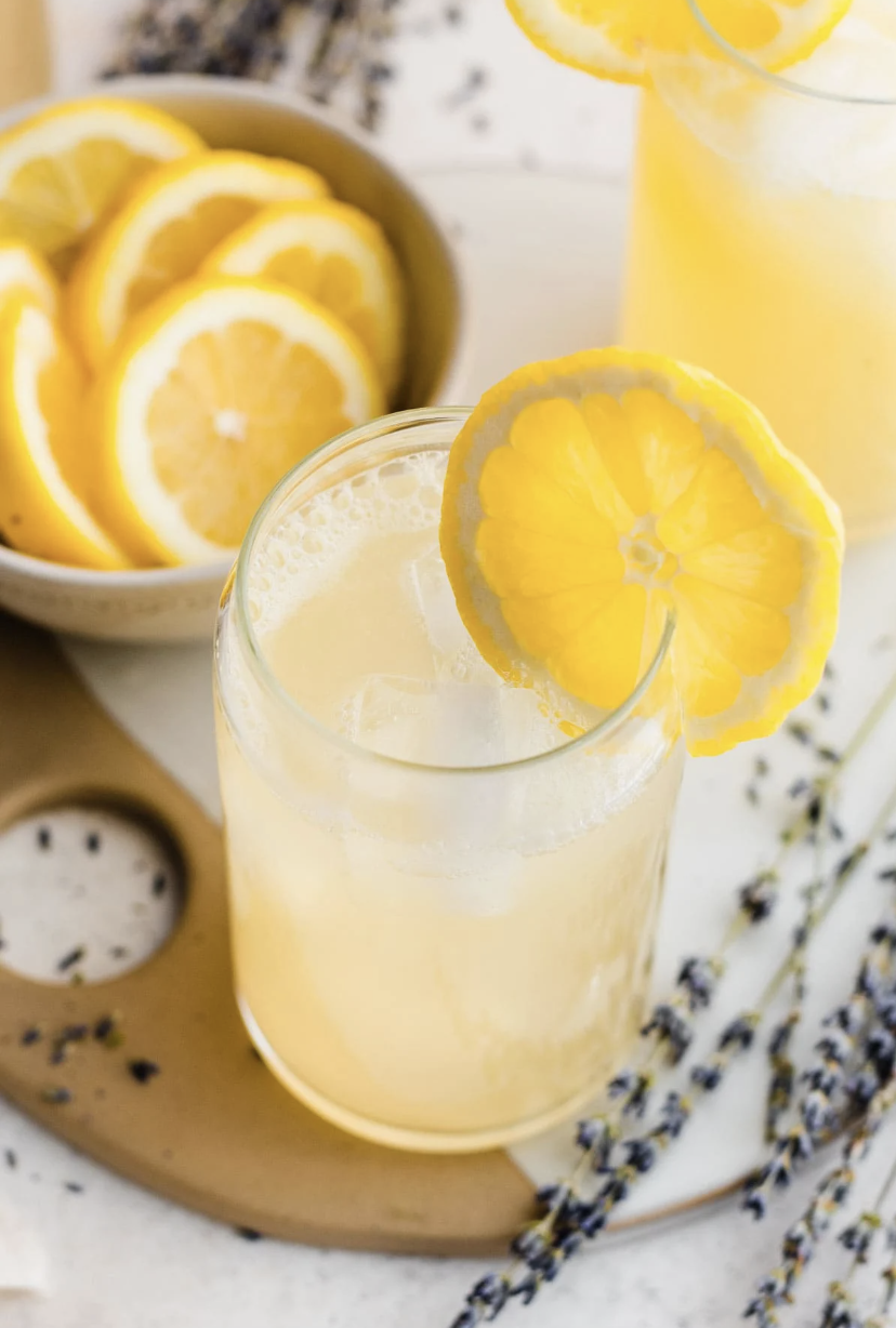 A glass of refreshing lemonade with ice and a lemon slice garnish, next to a bowl of lemon slices and sprigs of lavender in the background