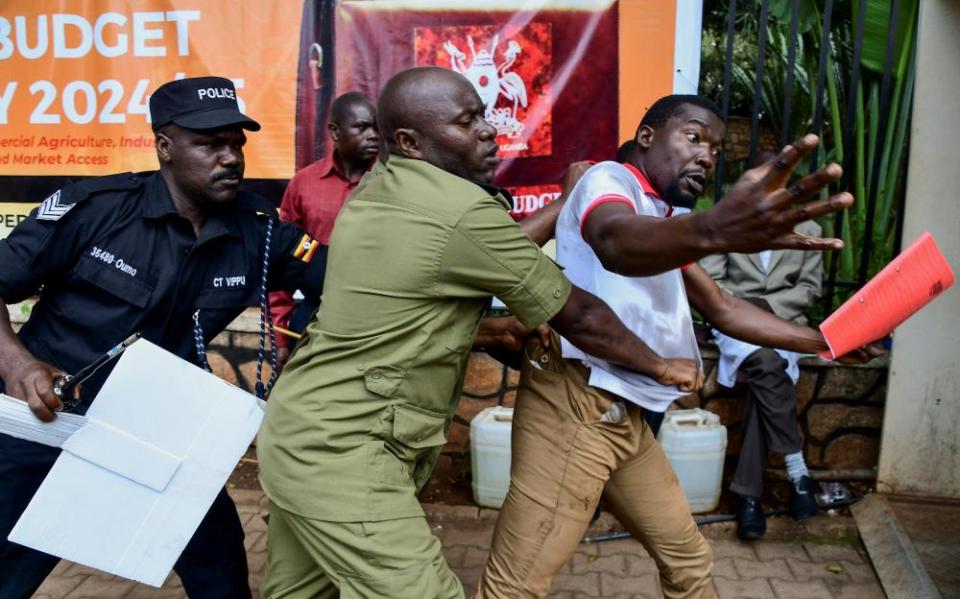 An African man wearing a white and red-T-shirt and brown pants, holding a red leaflet and with arms outstretched, is being pulled on from the back by a man in a black uniform and a man in an olive uniform.