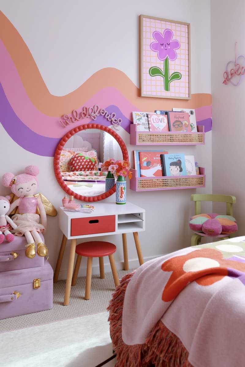 orange, pink, and purple squiggle line mural through middle of wall, round red mirror above small vanity desk, pink and red flower throw blanket, purple suitcase with dolls