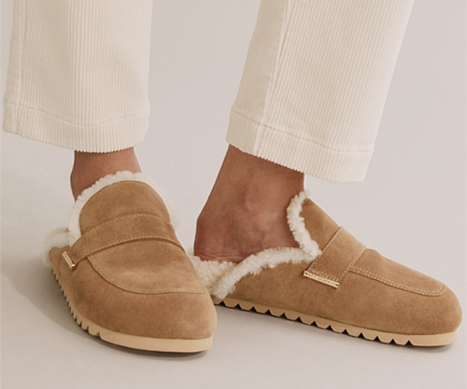 A close up of a person's feet inside beige colored suede mules lined with cream fluffy sheepskin.