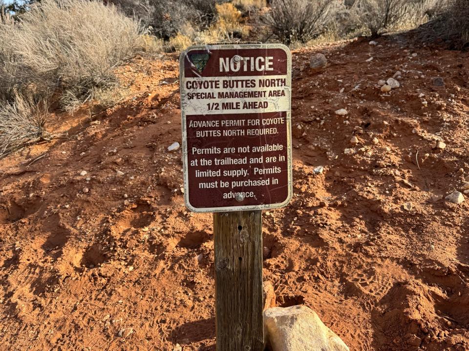 Brown and white safety notice sign by entrance to The Wave trail. The sign sits on a rocky hill with bushes in the background