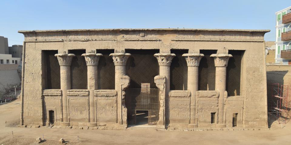 A picture shows the ouside of the temple of Esna on a sunny day. This rectangualar buildling's entrance is lined with columns.