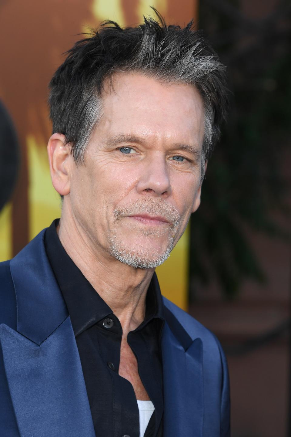 Kevin Bacon poses on a red carpet, wearing a dark suit with a white and black shirt underneath. He has short, styled hair and a slight smile
