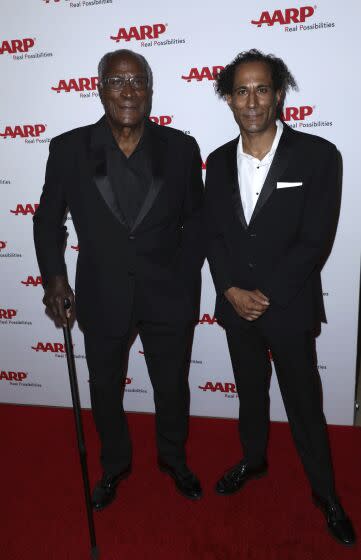 John Amos and K.C. Amos pose together on a red carpet in black formal attire.
