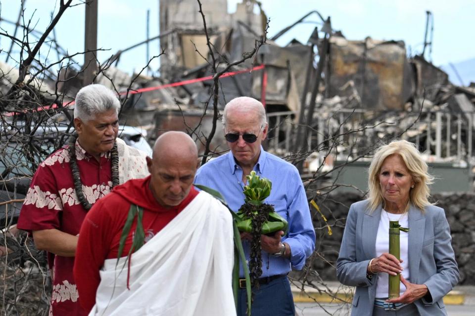 A man in blue shirt and sunglasses and a woman in gray jacket and white top, holding green items, follow two people