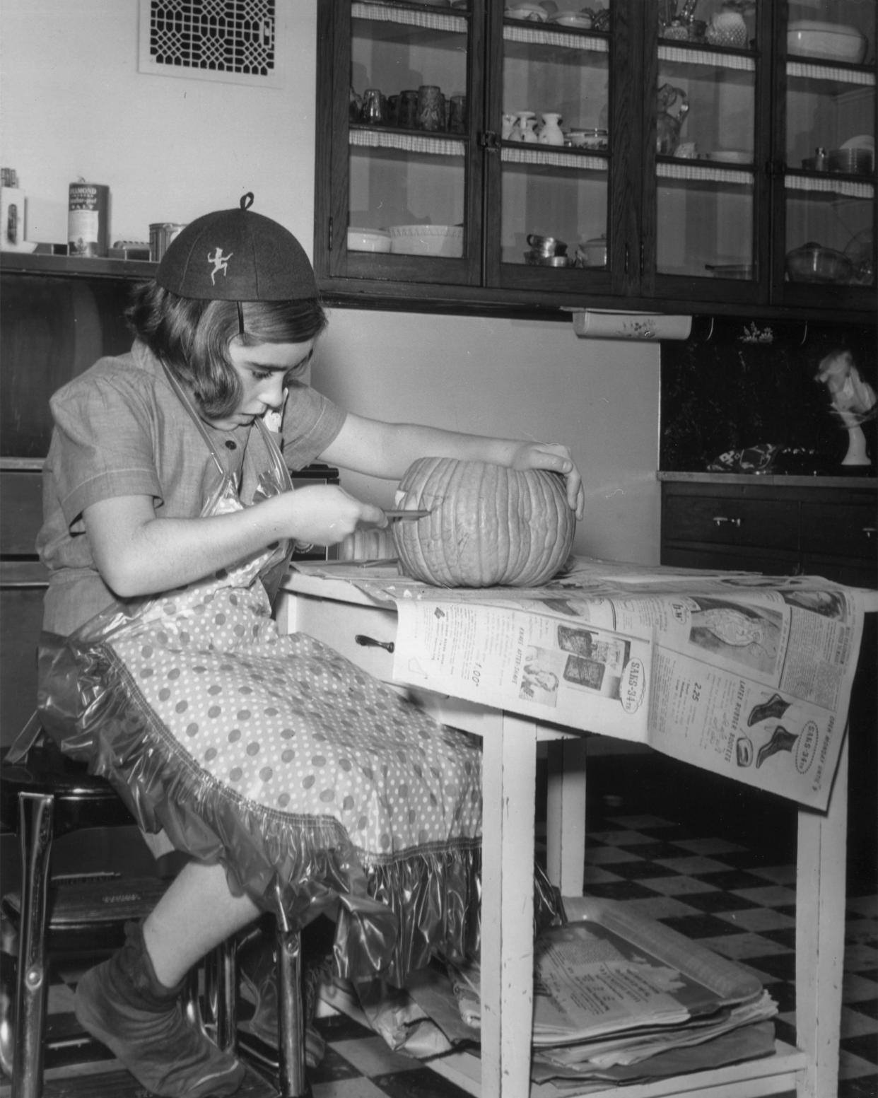 A young girl wearing a Brownie uniform and an apron carves a Halloween Jack-o-Lantern on a kitchen table.