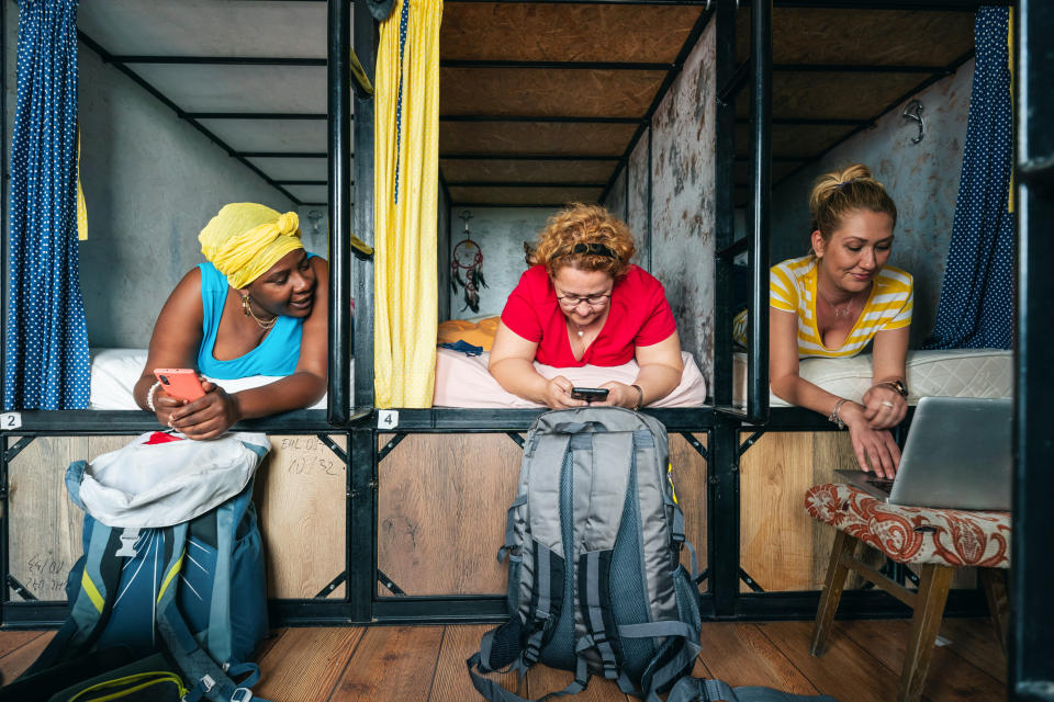 Three women relax in a hostel bunk bed area, using their phones and laptop. They have hiking backpacks beside them, suggesting travel or adventure