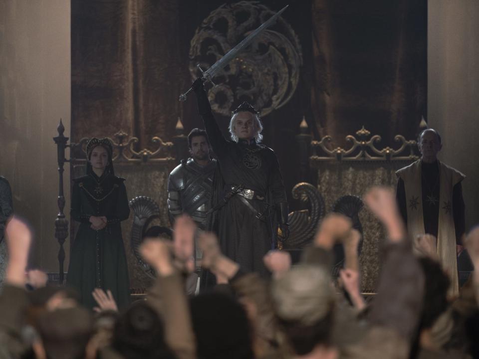 tom glynn-carney as aegon targaryen, standing in front of a crowd at his coronation and holding up a sword. behind him are his mother, wife, and ser criston cole, as well as a three headed golden dragon banner