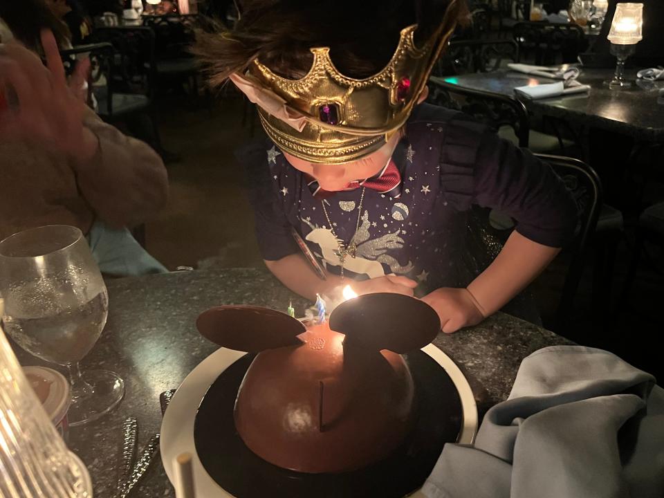 A little boy wearing a crown blows out candles on a Mickey Mouse cake.