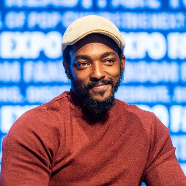 Anthony Mackie wearing a newsboy cap while he sits onstage at a fan expo