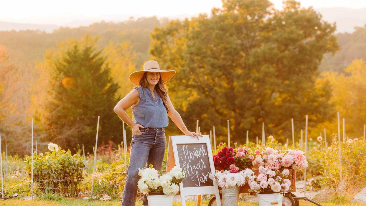 niki irving standing next to buckets of dahlias on her farm, wearing cropped jeans, flutter sleeve blue shirt, brimmed sunhat