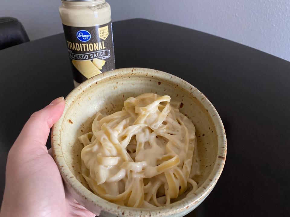The writer holds a bowl of Alfredo with Kroger-brand Alfredo sauce jar in background
