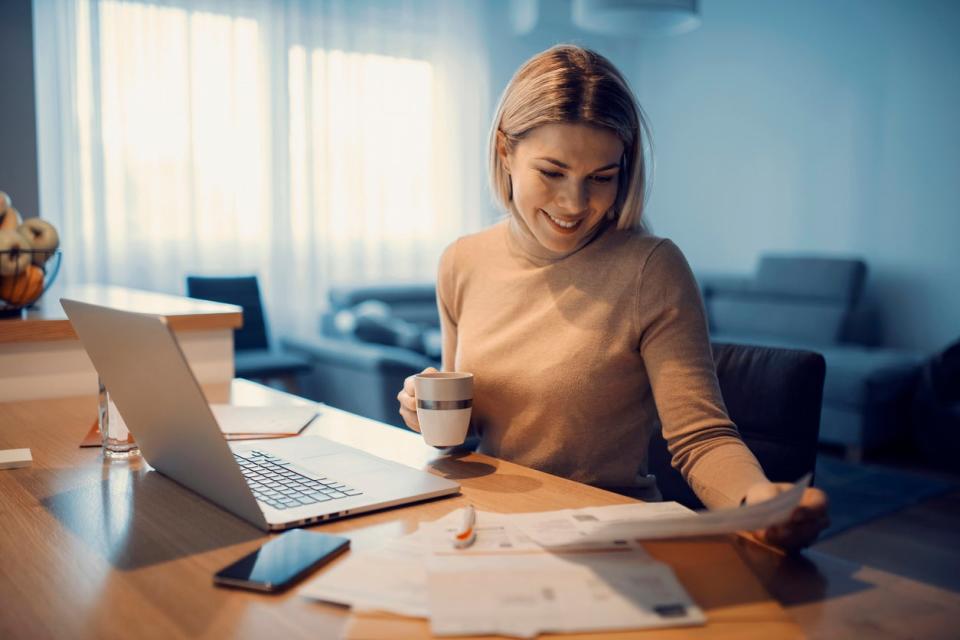 A woman smiles while looking at a document and holding a mug while sitting in front of a laptop. 