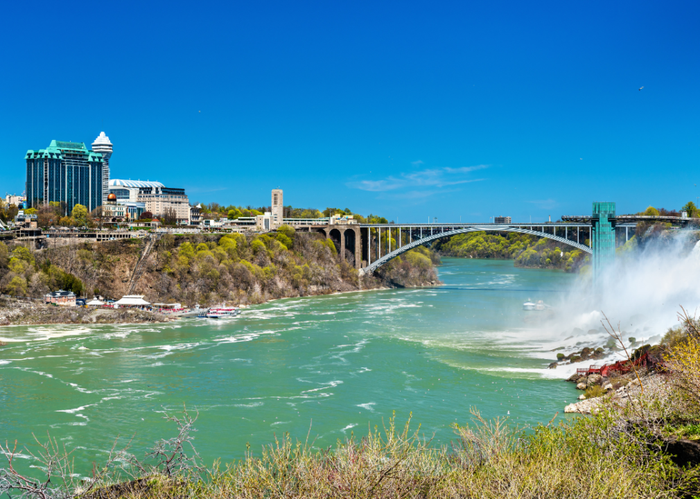 A steel arch bridge with suspension cables with river beneath and shoreline with trees and buildings. 