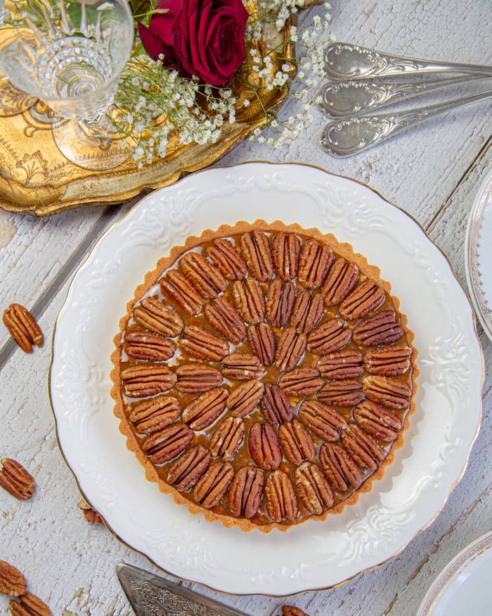 A pecan tart on a decorative white plate, surrounded by silverware, a red rose, and a crystal glass on a wooden table