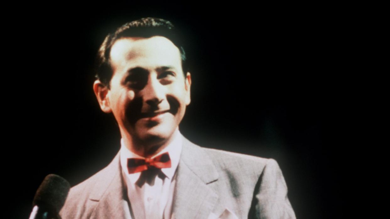 paul reubens as pee wee herman on a darkened stage, standing in front of a microphone and smiling
