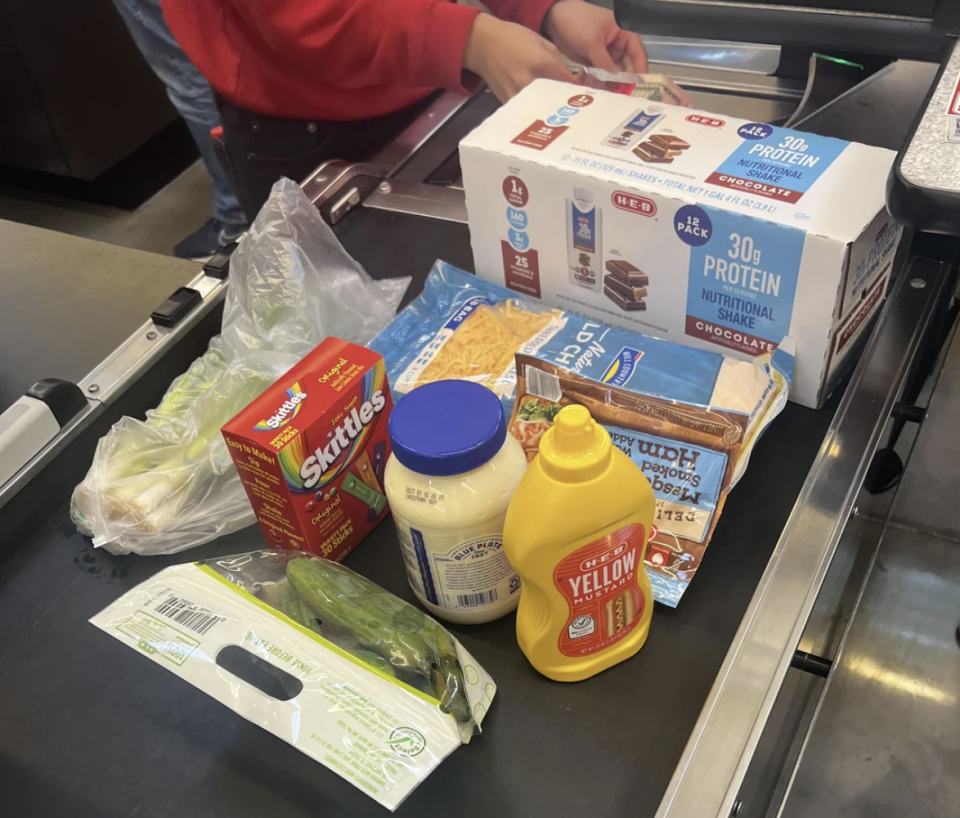 Grocery items on a checkout conveyor belt, including Skittles, mayonnaise, mustard, shredded cheese, cucumber, lettuce, protein shakes, and instant noodle soup