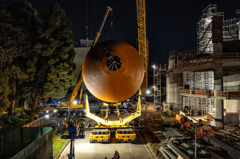 Front view of a horizontally aligned orange fuel tank, supported by a crane at night.