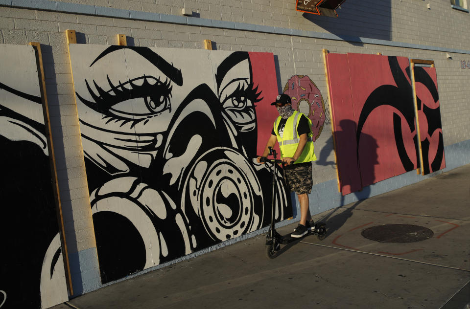 In this April 22, 2020, photo, a man in a mask rides a scooter by coronavirus inspired artwork on plywood covering the doors and windows of a temporarily shuttered donut shop in Las Vegas. (AP Photo/John Locher)