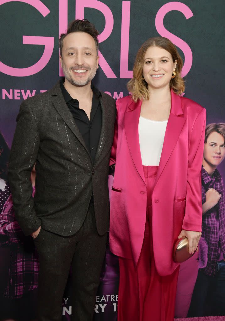 arturo perez jr and samantha jayne smile at the camera while standing in front of a pink and black movie poster for mean girls, he wears a black suit, she wears a pink suit