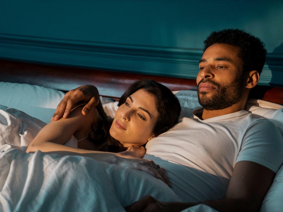 razane jammal and lloyd everitt as lyta and hector hall in the sandman. they're lying together in bed, tucked under the covers with lyta laying on hector's chest as warm sunlight breaks over them.