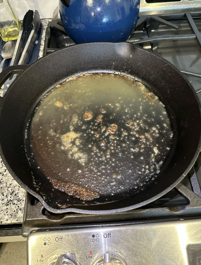 A cast iron skillet with oil on a stovetop, containing some food residue from previous cooking