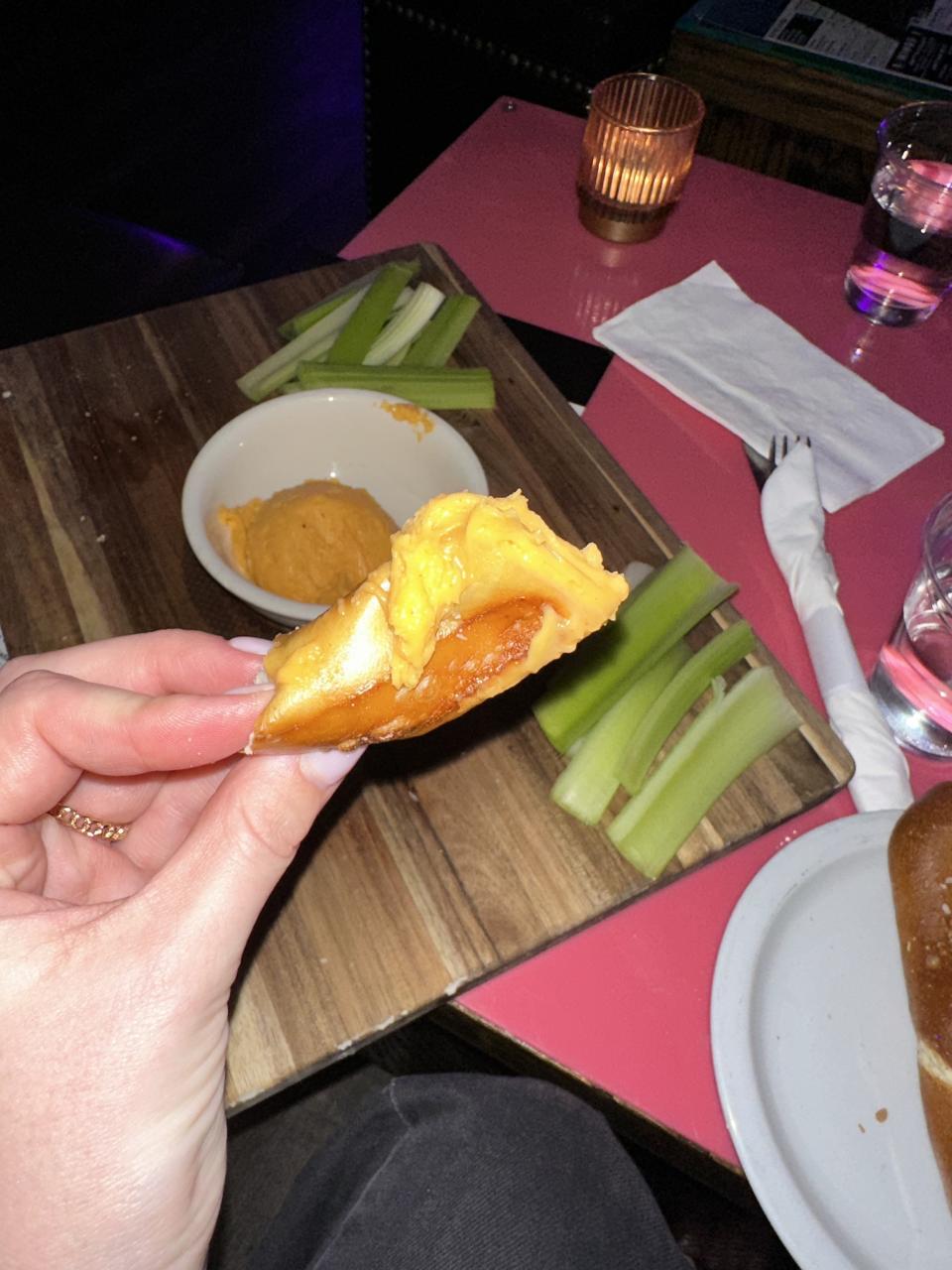 Person's hand holds a piece of buffalo chicken wing, with celery sticks and sauce on the side on a table