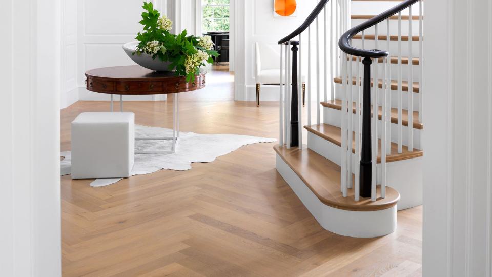 in an entry a grand freestanding curving staircase has natural wood steps, a white balustrade, and black bannisters, wood floor, an antique round table and a white leather cube