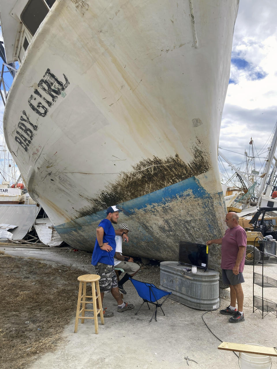 Shrimpers watch an NFL football game under the bow of the 75-foot-long trawler Baby Girl, which was grounded by Hurricane Ian, in Fort Myers Beach, Fla., on Sunday, Oct. 9, 2022. (AP Photo/Jay Reeves)