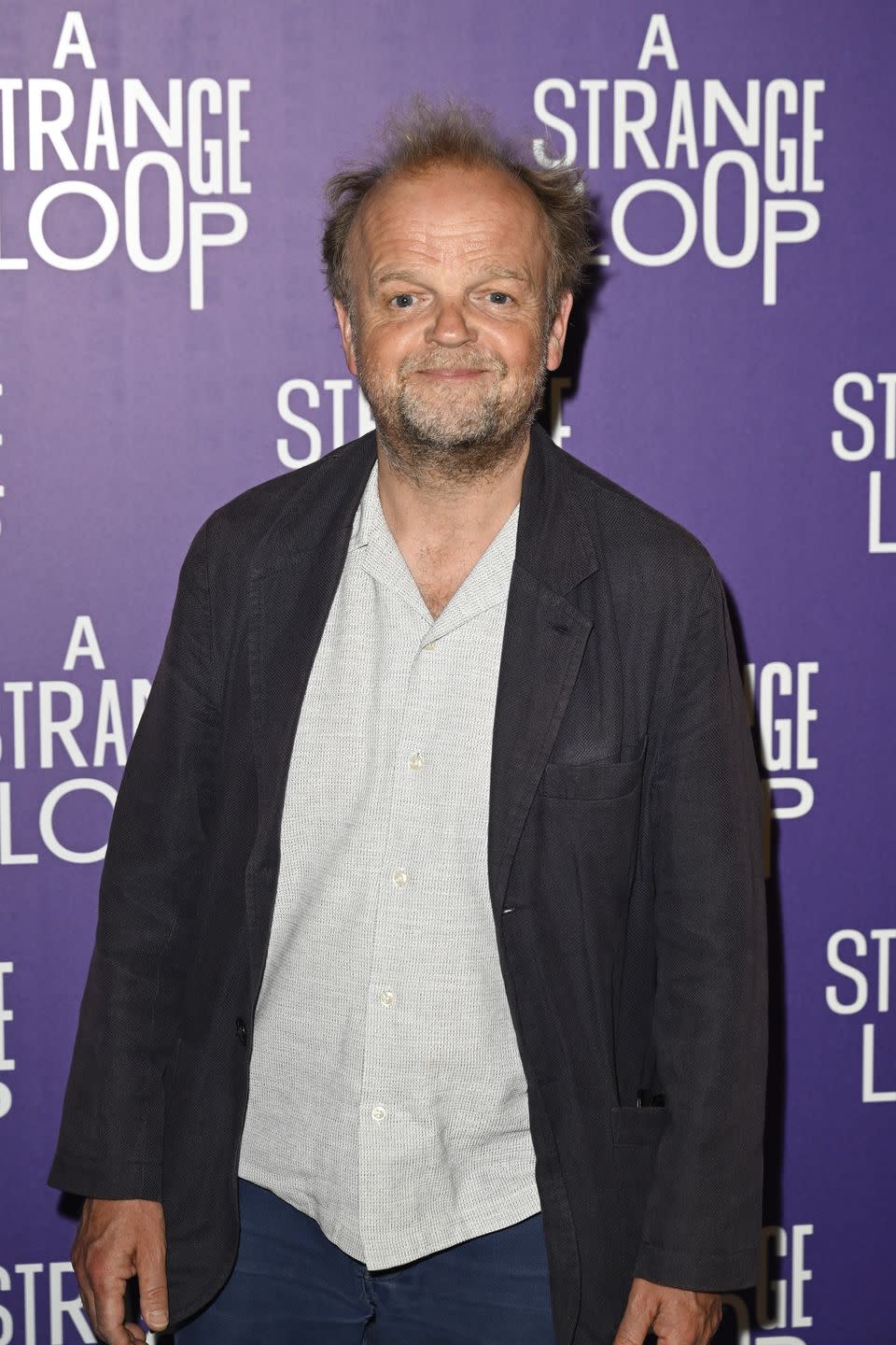 toby jones, an older man stands smiling at the camera, he has brown hair, wears a white shirt with jeans and black jacket