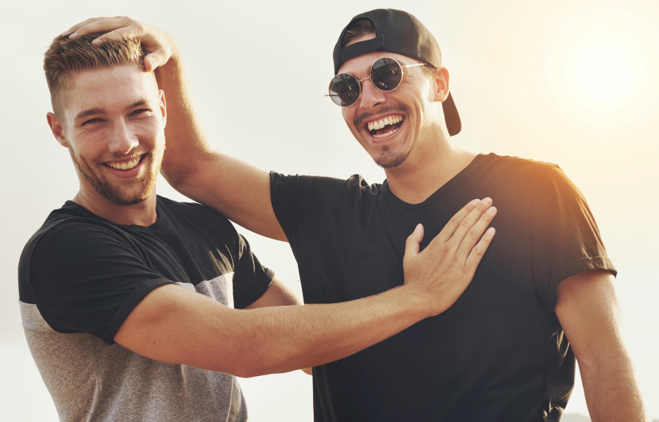 Two smiling men, one touching the other's head playfully. Both wearing casual t-shirts and sunglasses. Outdoors, sunlit background