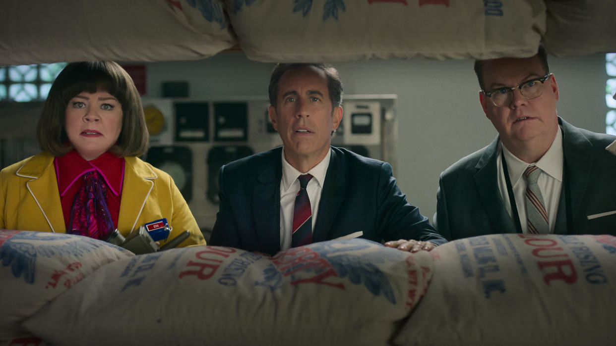  Melissa McCarthy, Jerry Seinfeld, and Jim Gaffigan looking shocked while standing behind a barricade of flour bags in Unfrosted. 