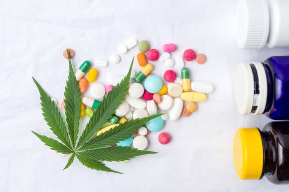 Overhead view of various types of pills lying on a surface with a marijuana leaf over them and three pill bottles lying on their sides adjacent to the pills.