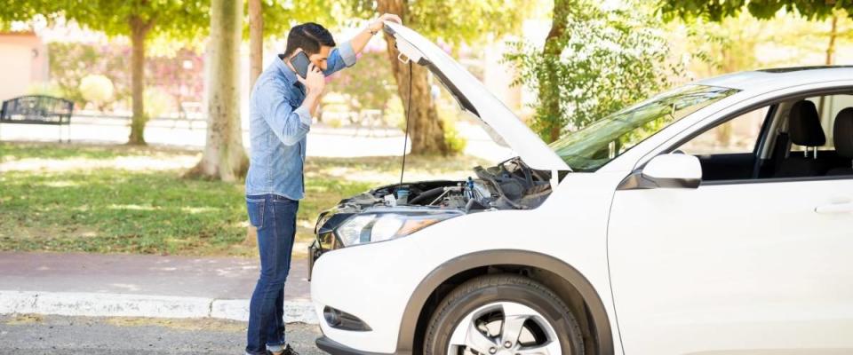 Young man standing in front of his broken car, looking at car engine and talking on cell phone