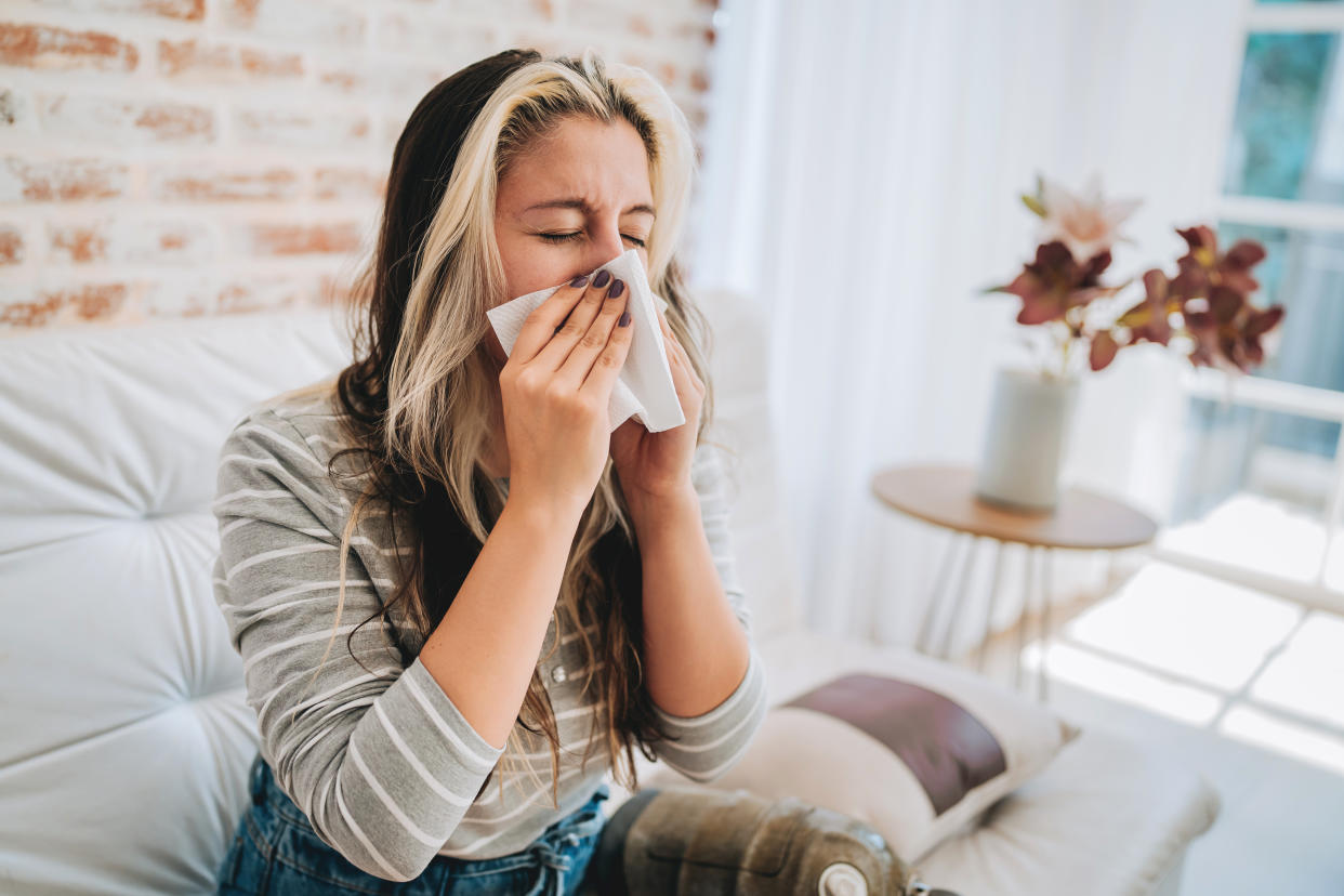 A woman holds a tissue to her nose as she sits on a couch.