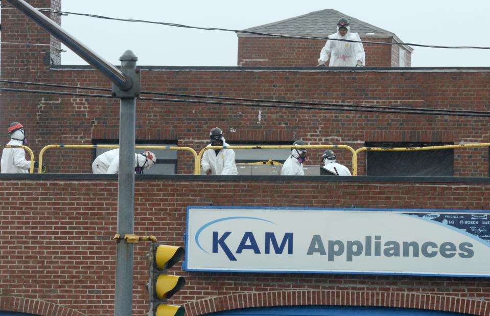 Dressed in hazmat suits, a work crew removes roofing material on April 6, 2022, atop the former KAM Appliances store that was torn town as part of a plan to reconstruct the Route 28 and Yarmouth Road intersection in Hyannis.