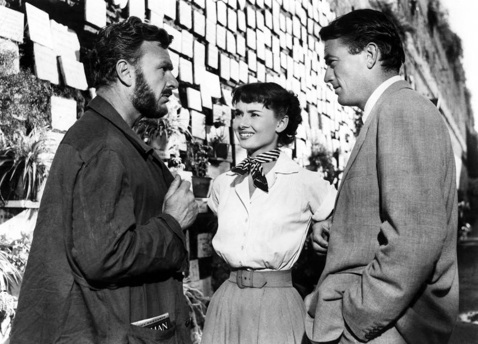 Gregory Peck, Audrey Hepburn, and Eddie Albert engage in conversation outdoors, with notes pinned on the wall behind them
