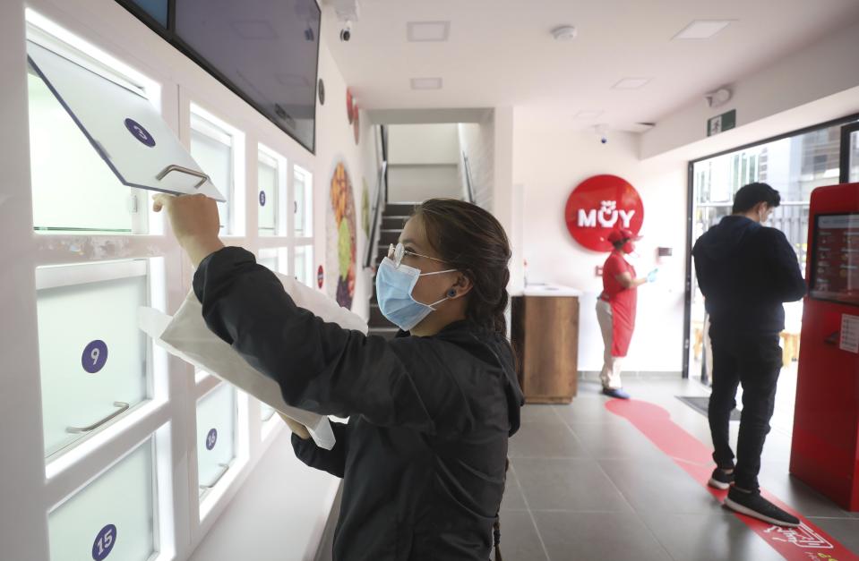 Una clienta recoge su pedido mientras otro coloca su orden usando una pantalla táctil en un restaurante de la cadena MUY de Bogotá el 13 de agosto del 2020. La cadena venía planificando la automatización de sus operaciones desde antes de la pandemia del coronavirus y ya abrió su primer local "sin contacto" físico entre personas. (AP Photo/Fernando Vergara) A customer picks up her food order from a locker while another places his order via touchscreen at a MUY restaurant, a Colombian fast food chain that is planning to turn its branches into automated restaurants, in Bogota, Colombia, Thursday, Aug. 13, 2020. The chain has opened its first "contactless store" in a commercial district of Bogota, where many restaurants have been forced to shut down because of a ban on sit-down dining. (AP Photo/Fernando Vergara)