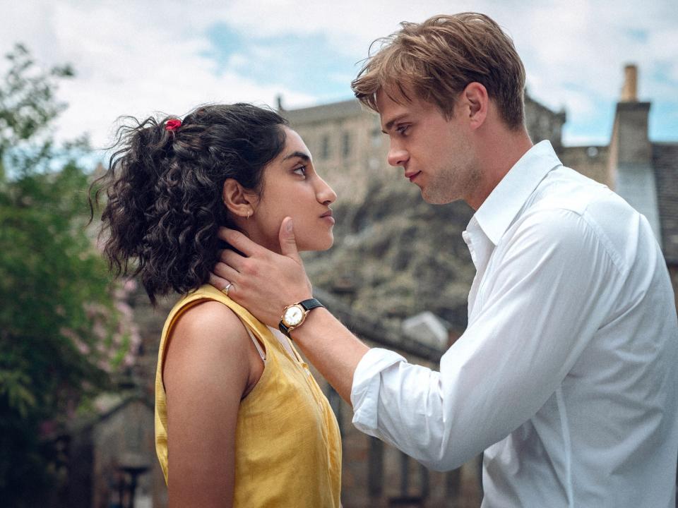 a young man cradles a woman's neck and jaw as they look intensely into each others' eyes face to face, standing outdoors in a picturesque village