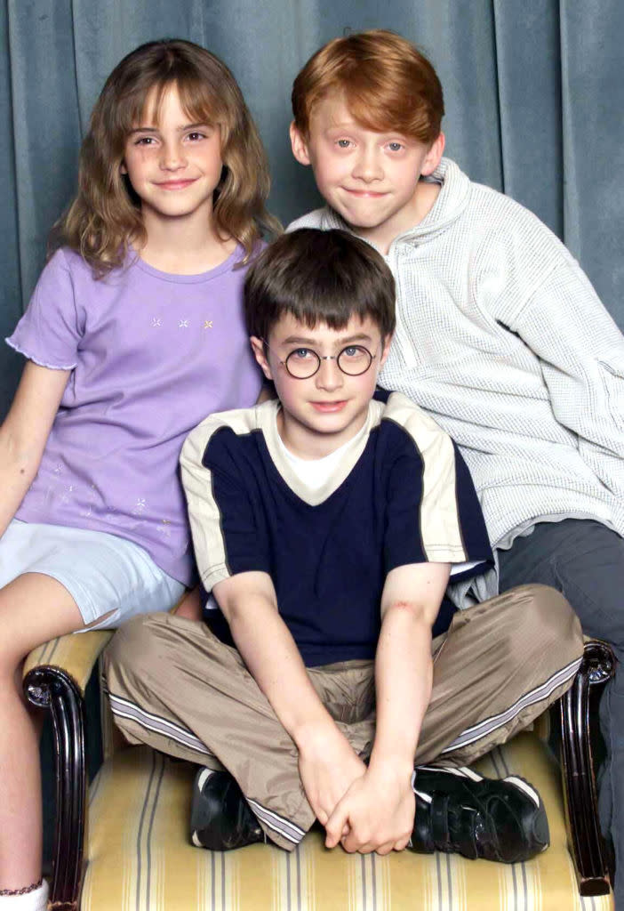 Emma Watson, Rupert Grint, and Daniel Radcliffe sit together, smiling, during a promotional event. Emma wears a casual outfit, Rupert a striped shirt, and Daniel a T-shirt
