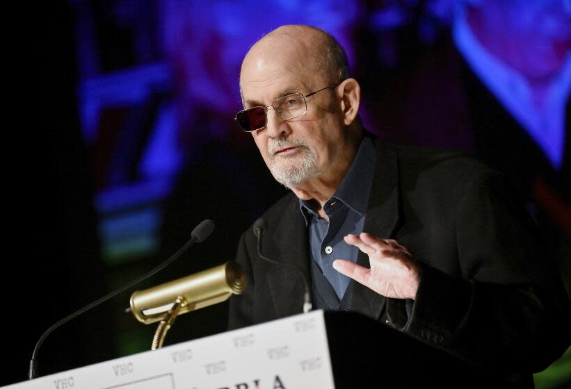 Salman Rushdie wearing glasses with one dark lens speaking at a podium with his left hand hovering