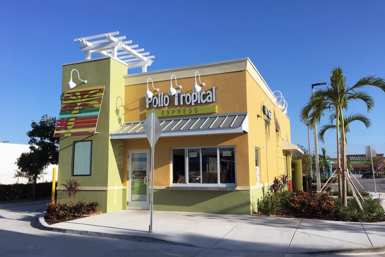 Front exterior of Pollo Tropical Express fast food restaurant, Miami, surrounded by sidewalk and lined by palm trees, with Walmart and parking lot in the background, against a clear blue sky