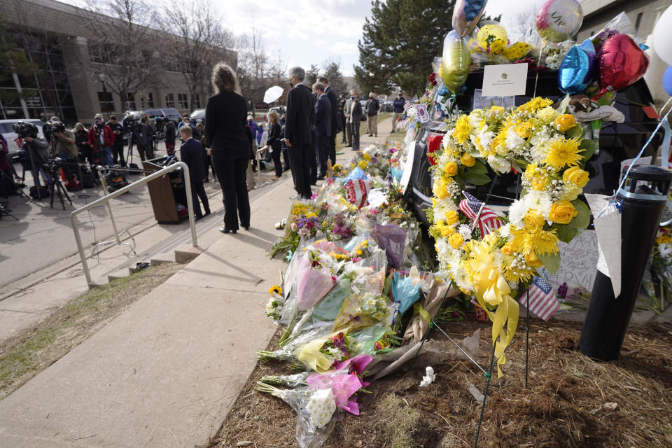 Wreaths and other tributes envelope a police cruiser as Boulder County, Colo., District Attorney Michael Dougherty, at the podium, makes a point to outline the ongoing investigation into the mass shooting at a King Soopers grocery store during a news conference outside police headquarters Friday, March 26, 2021, in Boulder, Colo. Ten people were killed in the shooting at the supermarket on Monday. (AP Photo/David Zalubowski)