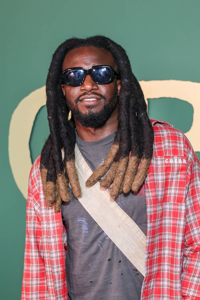 Man with long dreadlocks, wearing sunglasses, plaid shirt and shoulder bag at an event