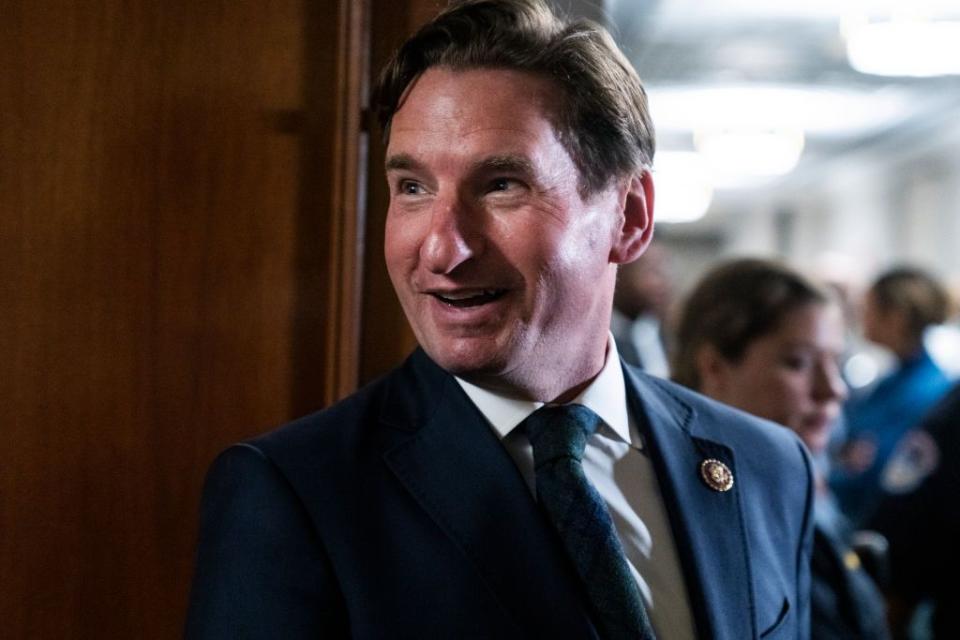 dean phillips smiles and looks left of the camera while standing in front of a wooden door, he wears a navy suit jacket with a pin on his lapel as well as a white collared shirt and blue and green tie