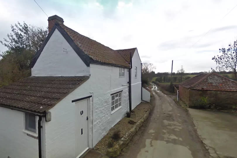 A white cottage stands on a country lane with fields in the distance.