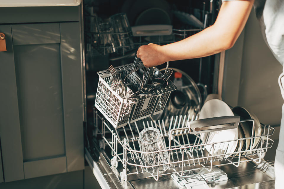 A person loads a dishwasher by placing a cutlery basket inside. The open dishwasher has various dishes and glasses