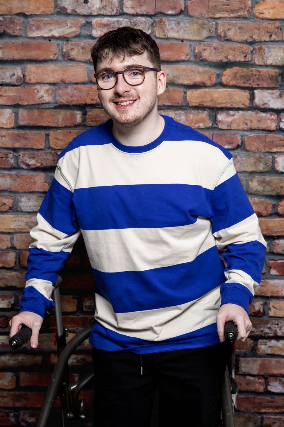 jack carroll as bobby in coronation street, a young man stands with a walking aid and smiles, he wears glasses and a blue and white striped top