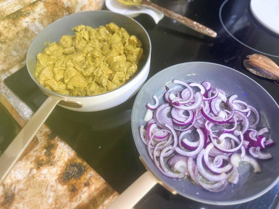 Two pans sitting next to each other on a black stovetop range. One pan holds chicken pieces in a yellow-brown marinade and the other holds slices of red onion